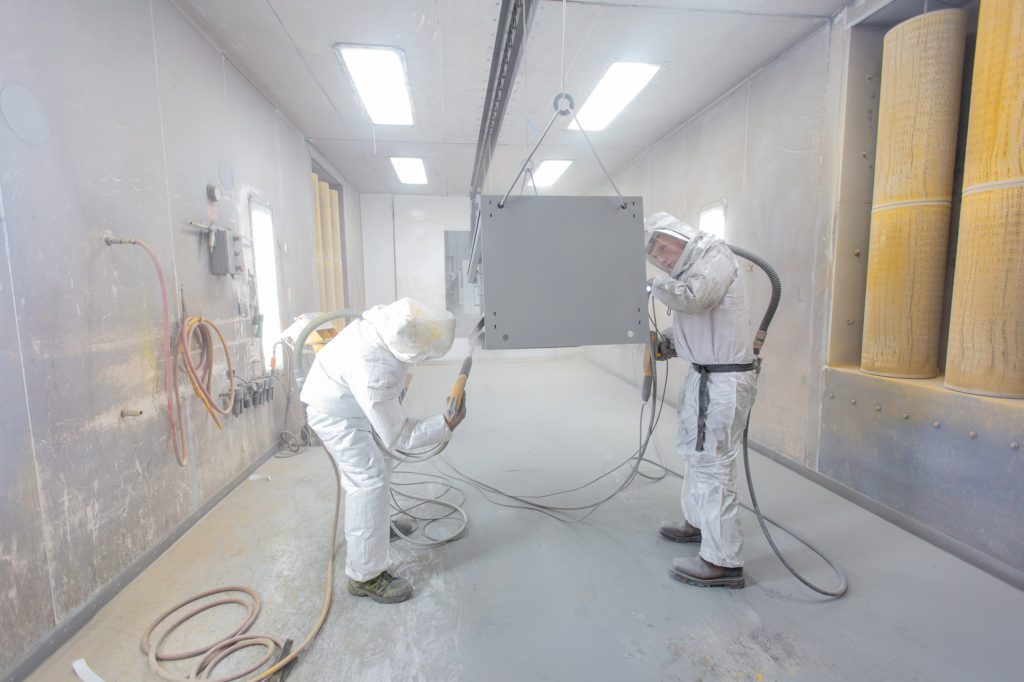 two men wearing protective gear completing powder coating on metal part
