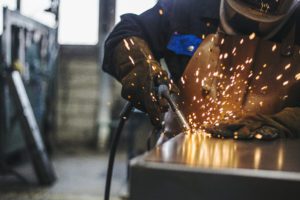 A welder fabricator holds a welding gun while fusing metals together. Sparks fly.