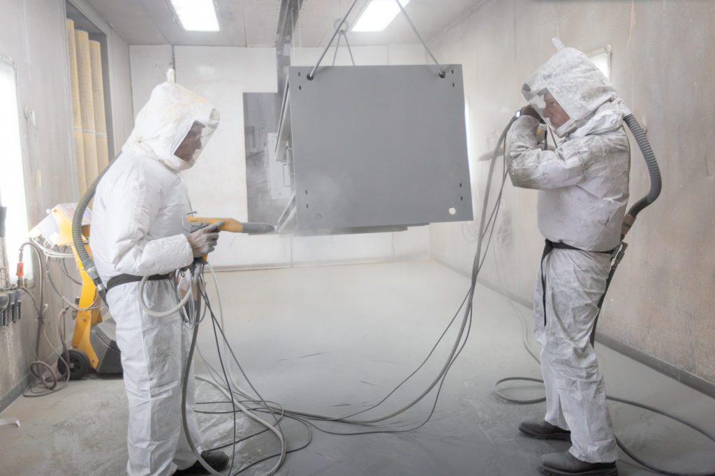 Two people in safety gear powder coating parts in the booth.