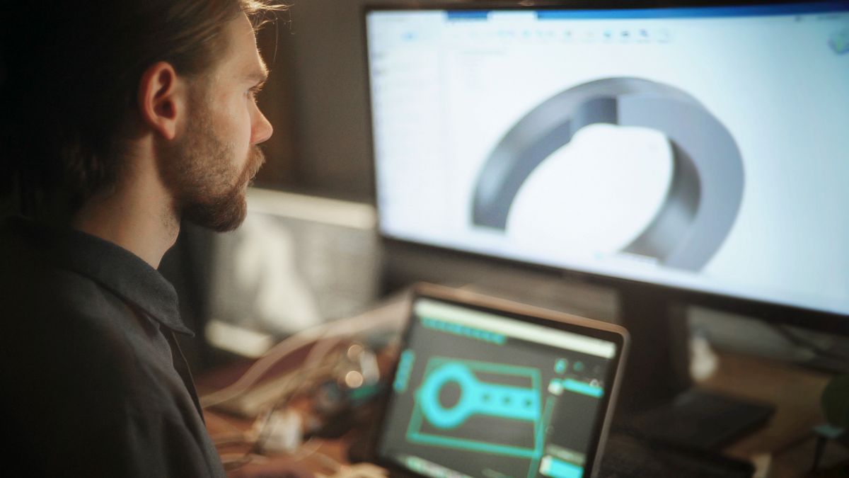 A bearded man looks at a metal component design on a computer.